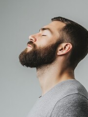 Obraz premium Side Profile of Young Handsome Man with Well-Groomed Beard Eyes Closed Relaxing Against Soft Grey Background Emphasizing Masculine Features