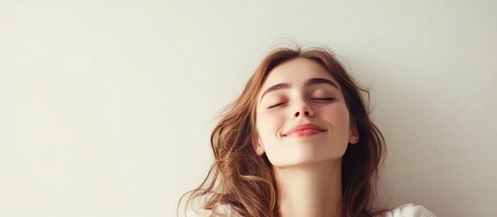 Smiling young woman with long wavy hair relaxing against a white wall, captured in a close-up shot emphasizing her peaceful expression and natural beauty.