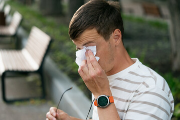 Young man wiping watery eyes with a tissue while holding glasses in a park, experiencing discomfort...