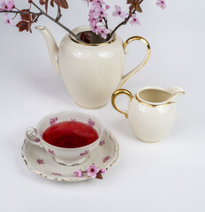Still life with a beautiful fine porcelain teacup, tea accessories and a pink blossoming branch in an elegant porcelain teapot on a white background