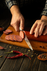 Hands expertly cut a delicious sausage into perfect slices on a rustic wooden board. Dried chilies...