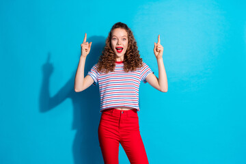 Joyful young woman in striped shirt and red pants pointing upwards against a vibrant blue background
