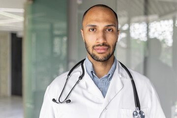 Close-up portrait of a doctor in a white medical coat, a man looks seriously and focused at the camera, the doctor works inside the clinic office at his workplace.