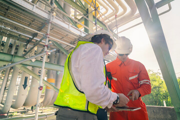 Two engineers in safety gear discussing project details using a tablet under a complex industrial pipeline structure, showcasing teamwork, modern technology, and operational expertise.