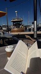 A relaxing harbor scene featuring a docked vintage boat, a book, and a takeaway coffee cup under sunny skies.
