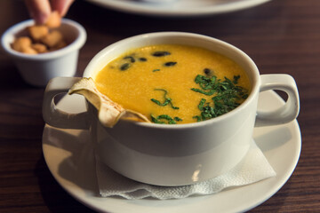 A cup of creamy pumpkin soup, hand taking croutons in background