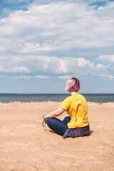 Young woman sits alone on a sandy beach under a serene sky with ocean waves