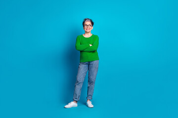Confident elderly woman with grey hair and glasses posing in green sweater against a vibrant blue background