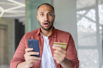 Portrait of disappointed and deceived man, holding bank credit card and phone in hands, account...