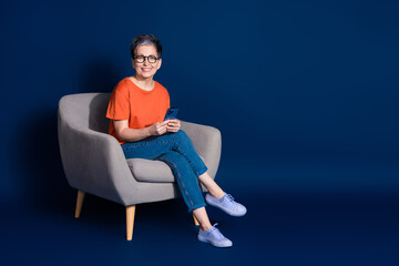 Full body photo of attractive senior woman sit armchair hold device dressed stylish orange clothes...