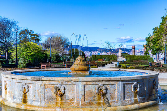 Stone fountain decorated with carved flowers, water jets coming out from central pipes, Joan Brossa Gardens on Montjuic, trees and cityscape in background, sunny day in Barcelona, ​​Catalonia, Spain