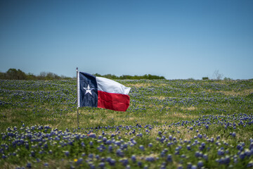 Texas Flag Blowing In The Wind In Rural Field of Bluebonnet Flowers With Blue Sky