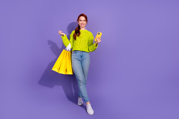 Cheerful young woman in green sweater holding shopping bags and smartphone over vibrant purple background