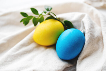 Easter background. Blue and yellow Easter eggs decorated with green twig on white fabric surface, close-up, copy space, soft focus