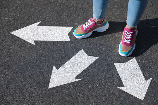 Woman standing near white arrows pointing in different directions on asphalt road, closeup. Concept of choice and making decisions