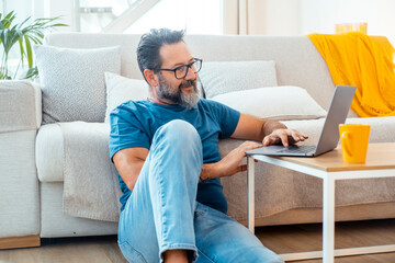 Man sitting comfortably on the floor in his apartment with a laptop placed on a small table, browsing the web to find interesting content, engaging with social media, and working remotely 