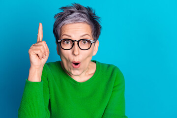 Surprised mature woman with glasses pointing up in green sweater on blue background