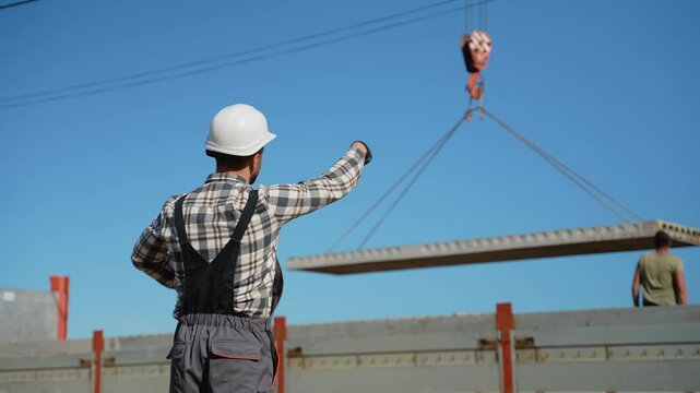 A foreman in a hard hat signals to a crane operator while lifting a concrete slab at a busy construction site