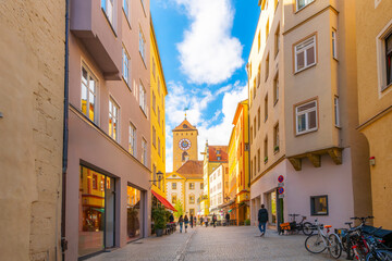 A cobblestone street with shops and sidewalk cafes with the Old Town Hall clock tower in the distance, in the Altstadt medieval old town of Regensburg, Germany.