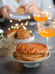 A plate of traditional French galette des rois or King cake, glasses of ciders, with person eating it in background with lighting bokeh. Cake made with puff pastry. It's usually served on Epiphany.