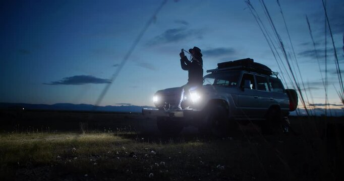 Young woman sitting in her car surrounded by nature at night, using her smartphone to photographing while embracing the peace and tranquility of the serene landscape