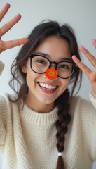 A cheerful young woman with long braided hair playfully poses with a bright flower on her nose, wearing stylish glasses and a cozy sweater. Her joyful expression radiates warmth and happiness