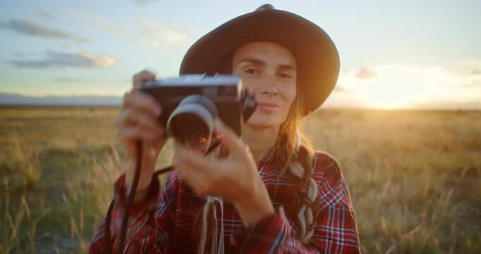 Young woman photographer taking pictures with vintage camera in a field at sunset. Cheerful Female traveler photographing on retro camera in a breathtaking landscape