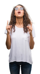 Naklejka premium Young hispanic woman wearing sunglasses amazed and surprised looking up and pointing with fingers and raised arms.