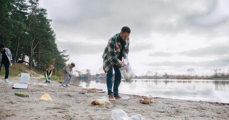 Scene portraying effort to raising awareness about impact of human waste on natural habitats. Male walking on sand beach while picking up pieces of plastic or other non organic material.