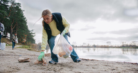 Camera focus on charming woman walking on beach with bag and collecting plastic from sand. Picking...