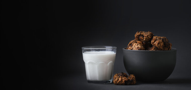 Oatmeal cookies in a black bowl near a glass of milk on the dark black background. Diet cookies with chocolate drops. Diet and healthy nutrition concept. Copy space for a free text