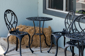 An empty black metal table with two wrought iron chairs on a patio. The building's wall is pale blue with grey trim. There's a sliding window with white trim. A bale of hay sits on the terrace.