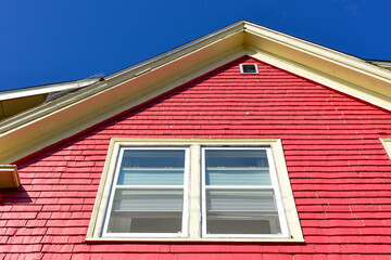 The middle roof peak of a vibrant red wooden cottage. The exterior wall of the house is covered in Cape Cod wood siding, two double-hung windows with cream trim and curtains. The sky is cloudy grey.