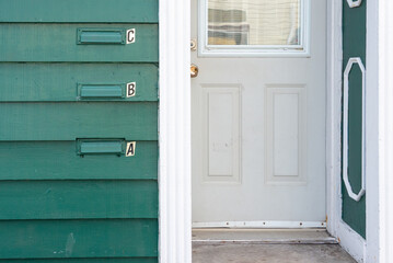 The exterior wall of a green wooden apartment building with three metal mail slots. There's a letter, a,b, and c, next to each letter slot. The apartment entrance door and trim are white colored.