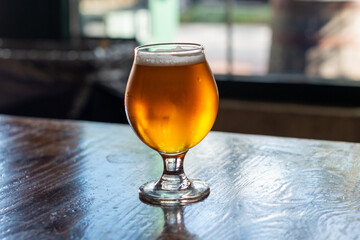 A tulip shaped clear beer glass filled with a cold blonde ale on a wooden table. The booze has a layer of froth on top of the liquor. The table is dark wood and there's a window in the background.