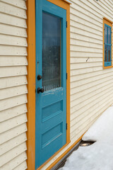A colorful teal blue color wooden panel door with yellow decorative trim on a white exterior wall. There's a long brass door handle on the wood door and a glass window on both sides of the door. 