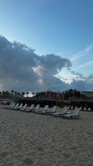 Obraz premium Empty sun loungers on a sandy beach with a dramatic sky, resort buildings, and palm trees in the background. Peaceful vacation scene with storm clouds and a tranquil seaside atmosphere