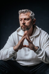 A close-up studio portrait of a mature man with salt-and-pepper hair and a serious, thoughtful expression. Wearing a white hoodie, he poses with hands clasped in front of him