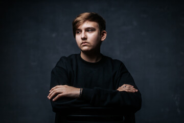 A young man in a black sweater sits with arms crossed, gazing thoughtfully into the distance. The...