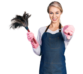 Beautiful young blonde woman wearing apron holding cleaning duster pointing to you and the camera with fingers, smiling positive and cheerful