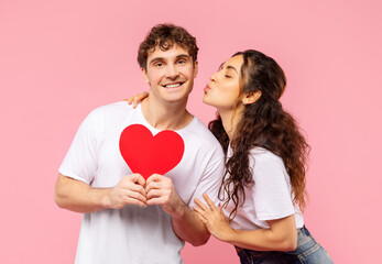 Valentine's holiday. Happy European guy holding red heart card, lady kissing him on cheek, standing over pink studio background. Love and romantic relationship