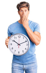 Young handsome man holding big clock covering mouth with hand, shocked and afraid for mistake....