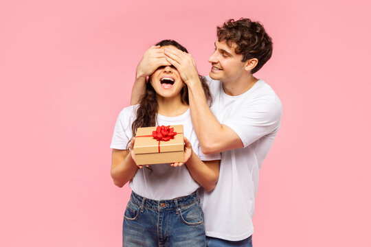 Happy man closing eyes of his surprised woman, making her surprise and giving gift box against pink background. Valentine's Day celebration