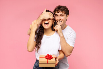 Happy guy closing eyes to wife, giving gift box and making shh silence sign over pink studio...