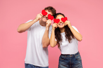 Lovers blinded by their love. Happy couple holding red heart-shaped cards over eyes and smiling, standing over pink background. St. Valentines Day concept