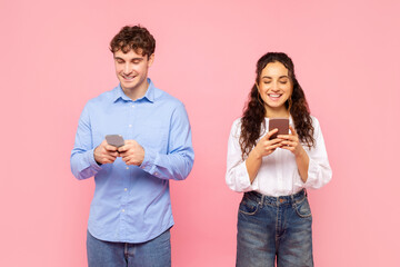 Cheerful man and woman holding their smartphones and chatting online, using app and smiling isolated on pink studio wall background