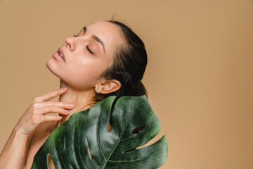 Beautiful young woman girl model with clean fresh skin touching her face in tropical green leaf closed eyes posing standing isolated in beige background . Cosmetology beauty and spa care concept.