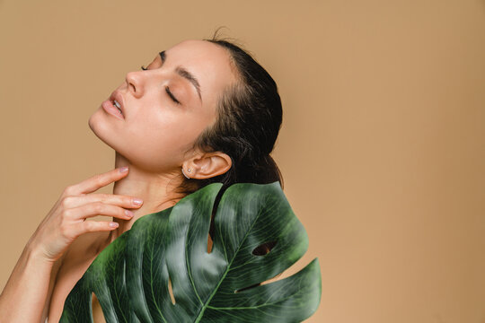 Beautiful young woman girl model with clean fresh skin touching her face in tropical green leaf closed eyes posing standing isolated in beige background . Cosmetology beauty and spa care concept.