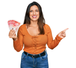 Beautiful hispanic woman holding 20 israel shekels banknotes smiling happy pointing with hand and finger to the side