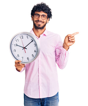 Handsome young man with curly hair and bear holding big clock smiling happy pointing with hand and finger to the side